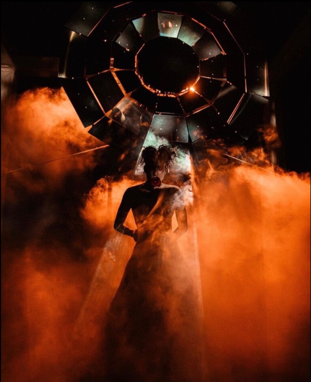 A cinematic ceremonial production at night, silhouette lit by fire and ambient smoke.